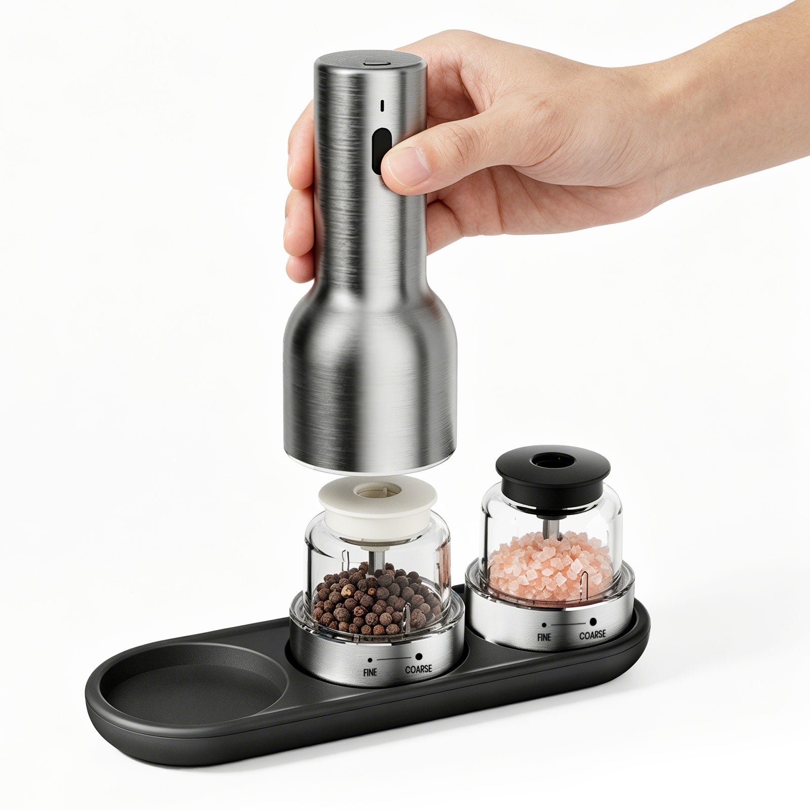 Hand holding a silver pepper grinder above a black tray with two glass containers filled with spices on a white background.